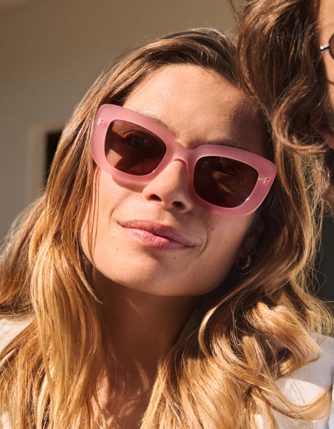 Primo piano di una donna sorridente con lunghi capelli biondi che indossa occhiali da sole rosa oversize.