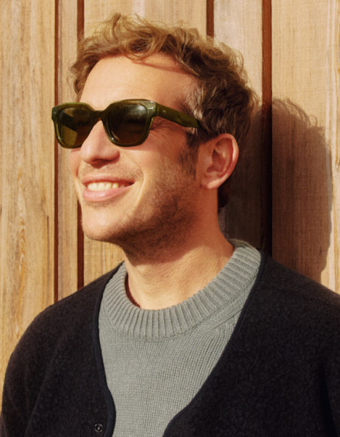 Uomo sorridente con riccioli corti, occhiali da sole verdi e maglione grigio, appoggiato a una parete di legno illuminata dal sole.