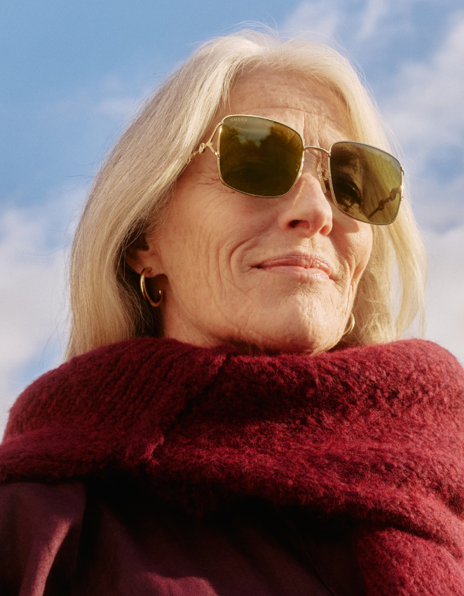 Donna dai capelli argentati con occhiali da sole e una morbida sciarpa bordeaux che guarda verso l'alto contro il cielo blu.
