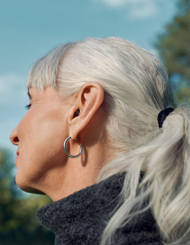 Profil einer Person mit langen weißen Haaren, die einen Creolen-Ohrring und ein schwarzes Oberteil trägt, vor einem unscharfen Hintergrund im Freien.