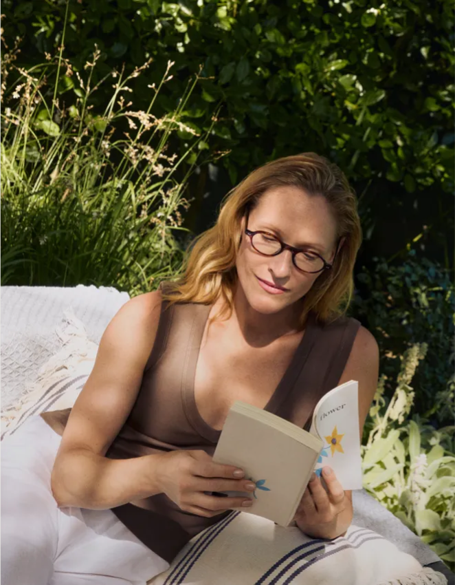 Eine Frau mit Brille liest an einem sonnigen Tag im Freien, umgeben von üppigem Grün, ein Buch.