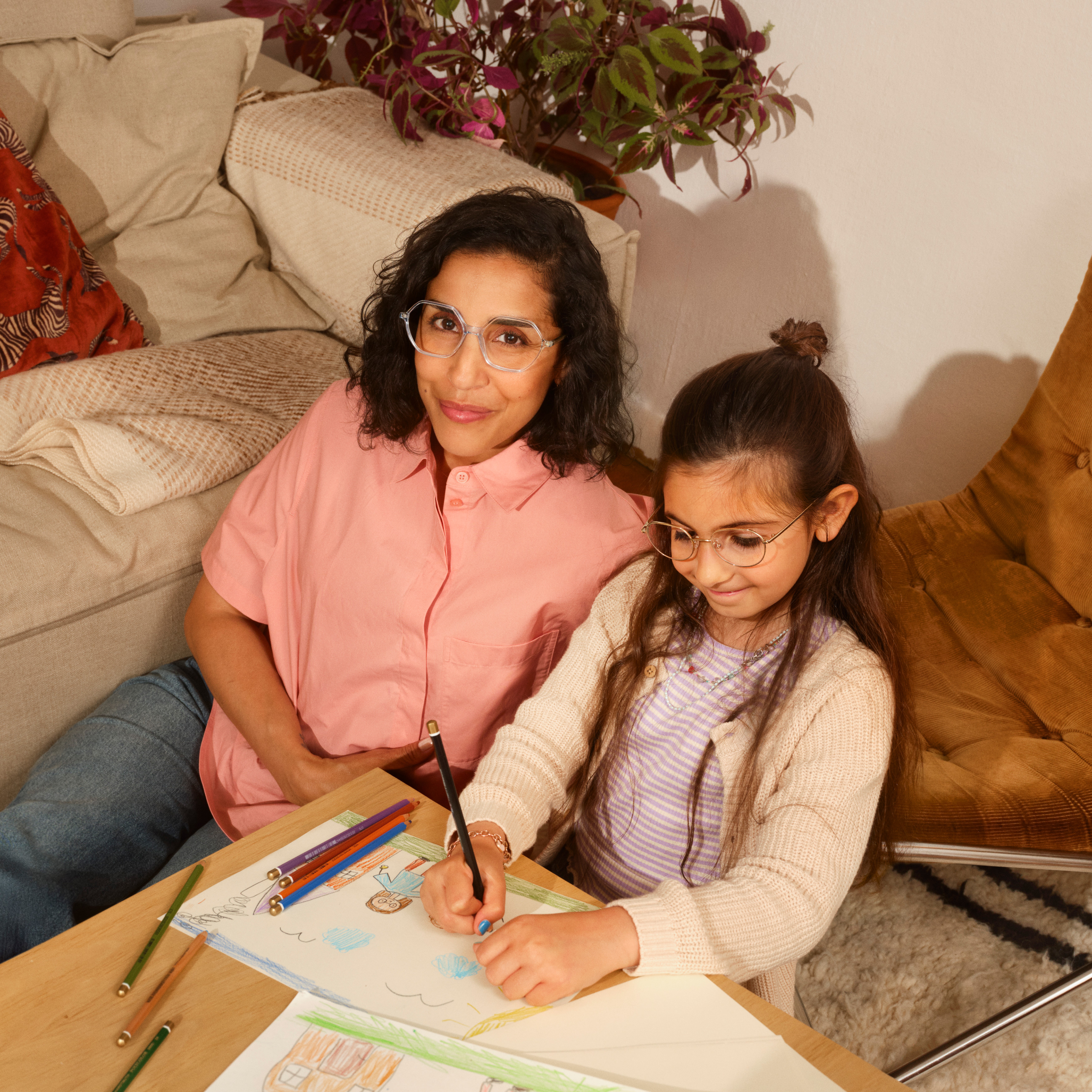 Eine Frau und ein Mädchen zeichnen gemeinsam an einem kleinen Tisch, umgeben von Buntstiften, in einer gemütlichen Wohnzimmeratmosphäre.
