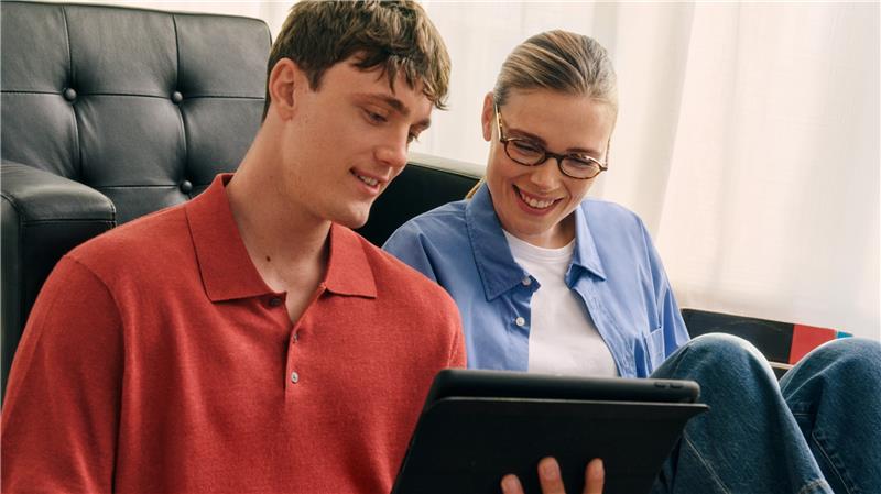 Ein Mann und eine Frau sitzen auf einem Sofa, lächeln und schauen gemeinsam auf ein Tablet in einem gut beleuchteten Raum.