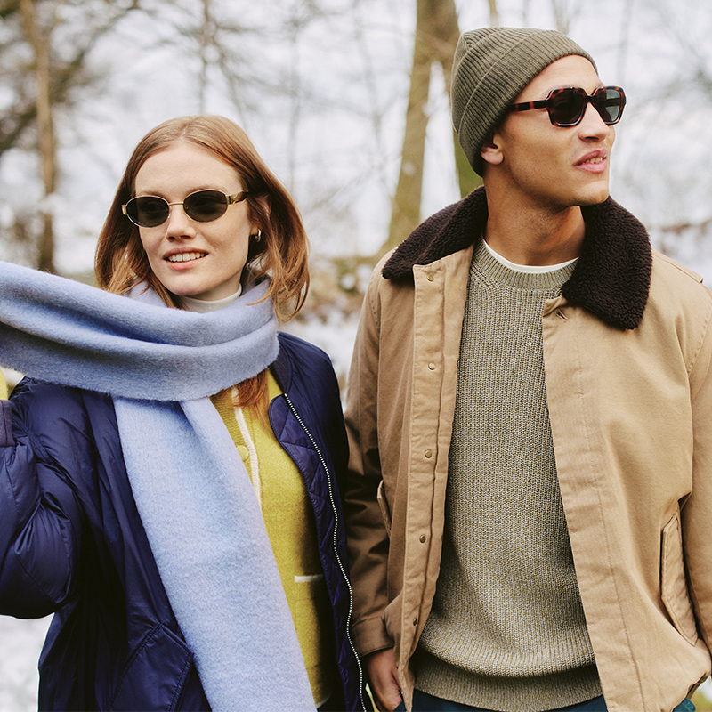 Ein lächelndes Paar mit Sonnenbrillen und Winterkleidung, darunter ein blauer Schal und eine Mütze, spaziert an einem bewölkten Tag im Freien.