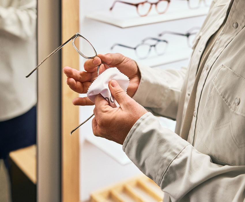 Eine Person reinigt eine Brille mit einem Tuch und steht dabei neben einer Präsentation mit verschiedenen Brillenmodellen.
