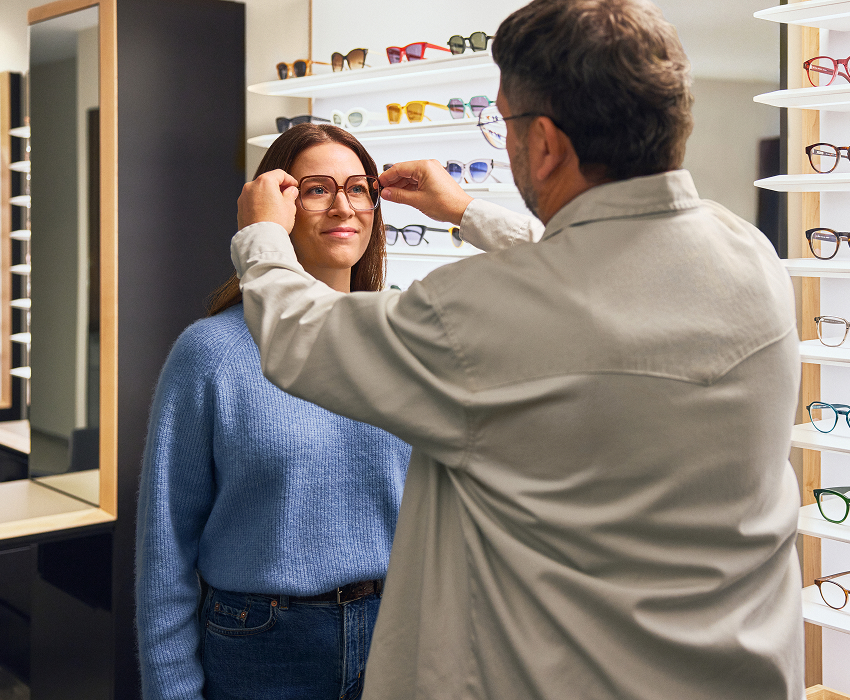 Ein Optiker hilft einer lächelnden Frau in einem Brillengeschäft beim Anprobieren einer Brille. Im Hintergrund sind Regale mit verschiedenen Brillenfassungen zu sehen.