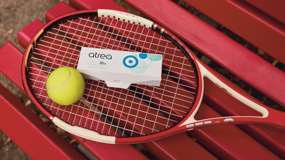 Una racchetta da tennis con una pallina gialla e una scatola con la scritta «atrea» su una panchina rossa all'aperto.