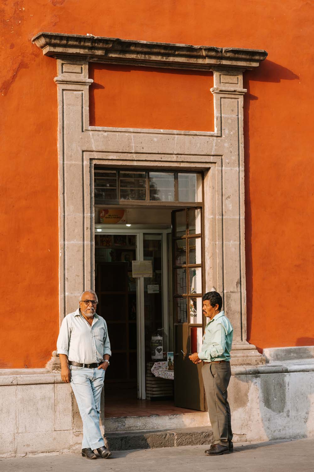 Two men stand outside a doorway of a red building. One leans against the frame, while the other looks down at a book.