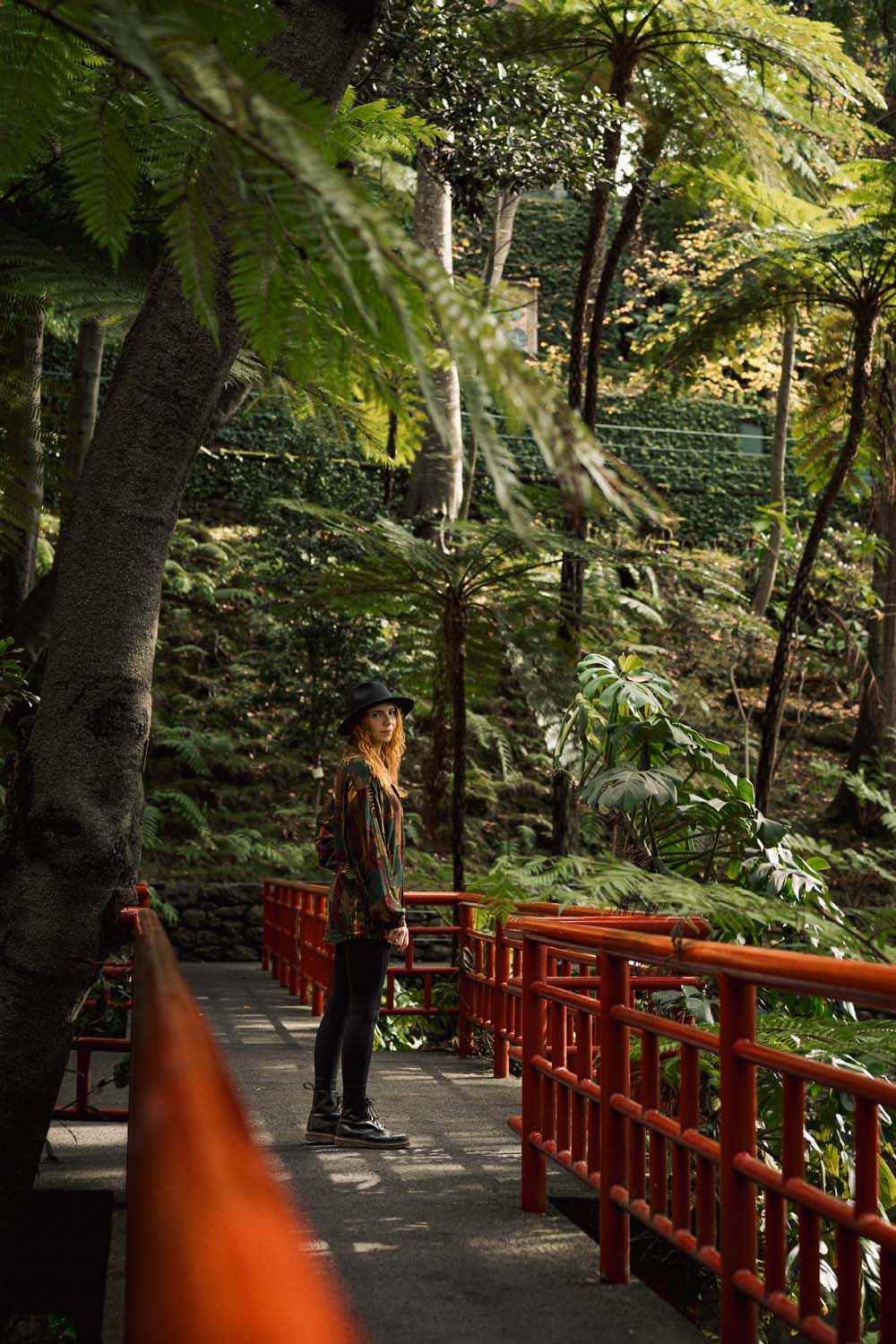 A person stands on a red bridge in a lush, green forest, surrounded by tall trees and ferns, wearing a hat and casual clothes.