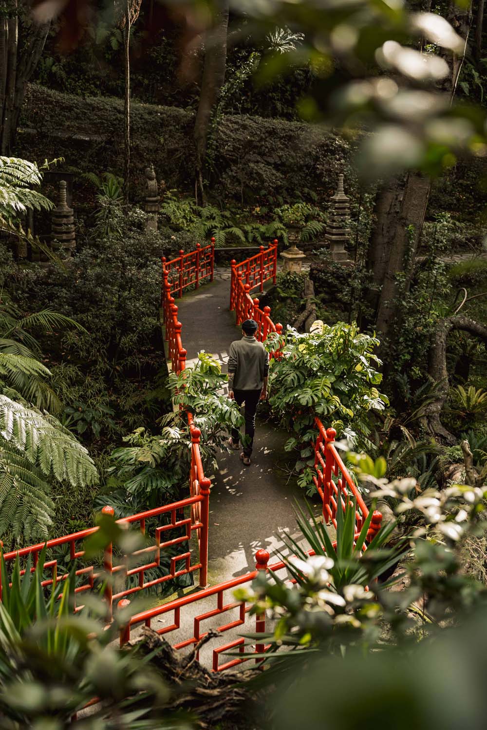 A person walks on a path with vibrant red railings surrounded by lush greenery in a serene garden setting.