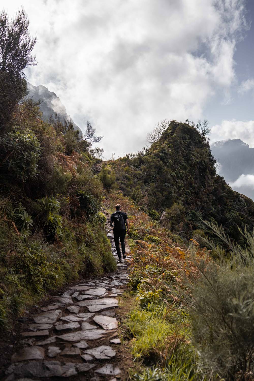 A person hikes up a stone path surrounded by lush greenery and hills under a cloudy sky.