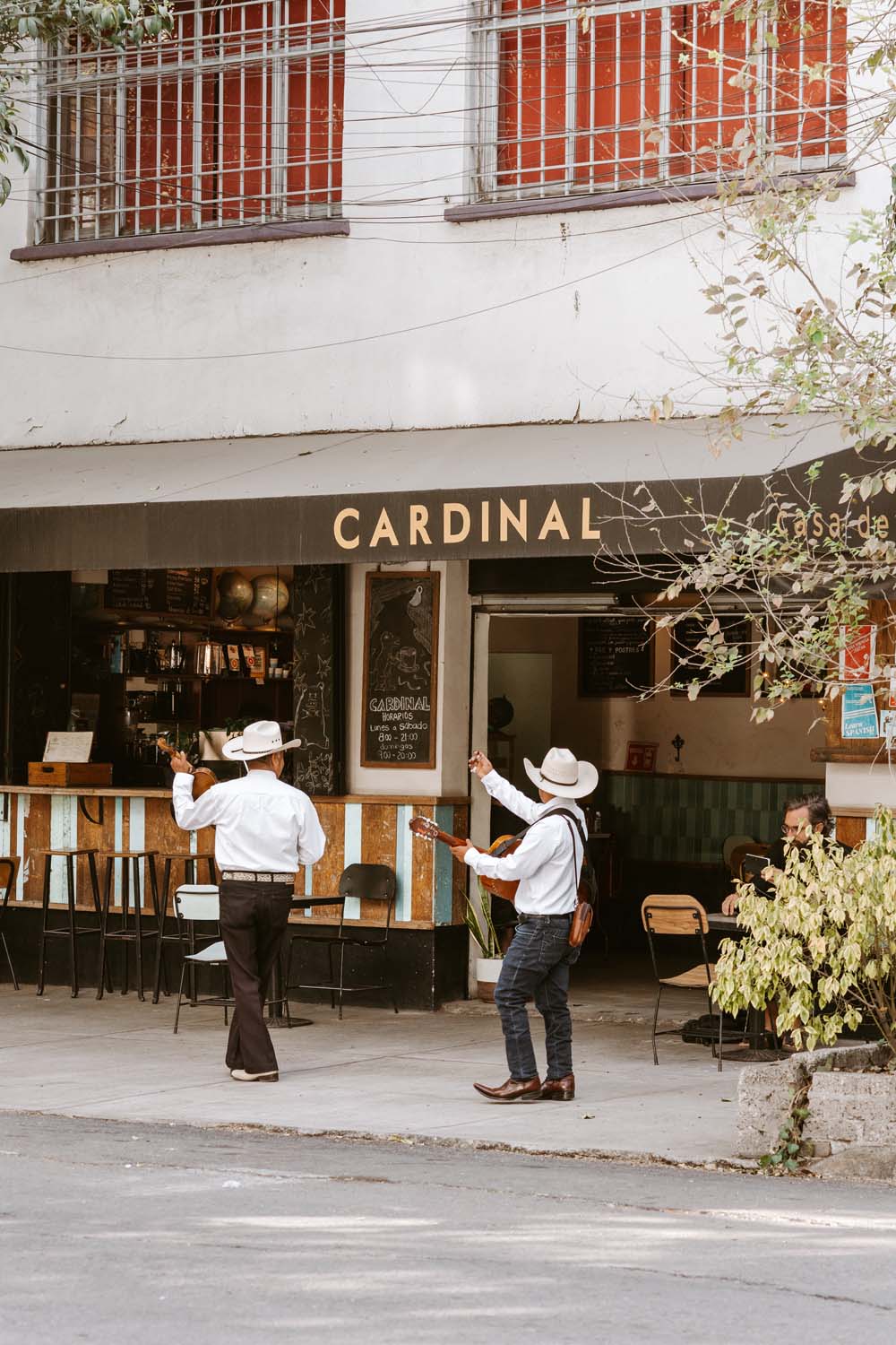 Two men in cowboy hats dance on the sidewalk in front of a cafe named "Cardinal." Another person sits nearby, watching.