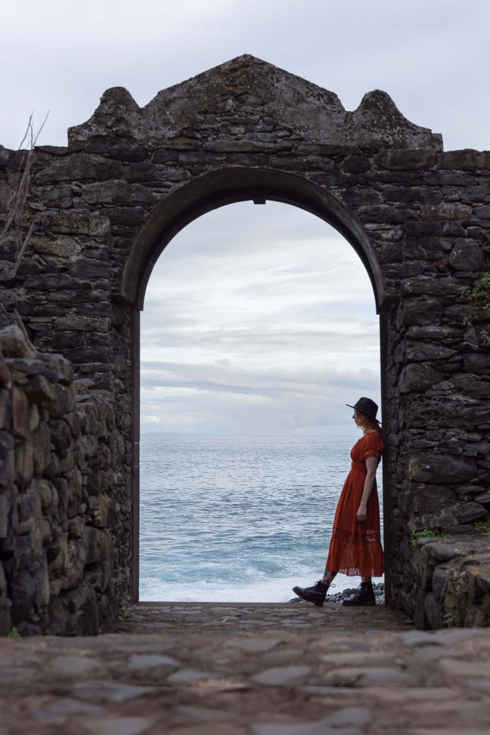 Person in a red dress and hat stands under a stone archway, overlooking a cloudy seascape.