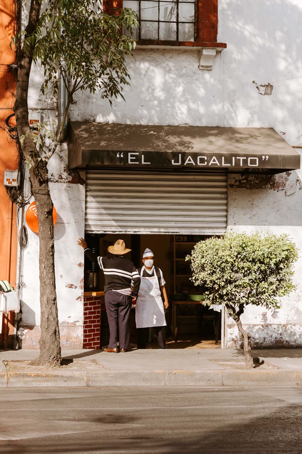 Two people stand outside a small shop with a sign reading "El Jacalito." One wears an apron and mask, and the other a hat. A tree is nearby.