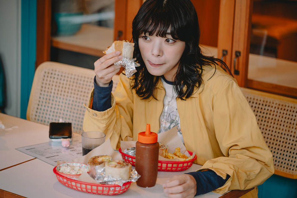 A person in a yellow jacket sits at a table eating a sandwich, with two red baskets containing food and a phone nearby.
