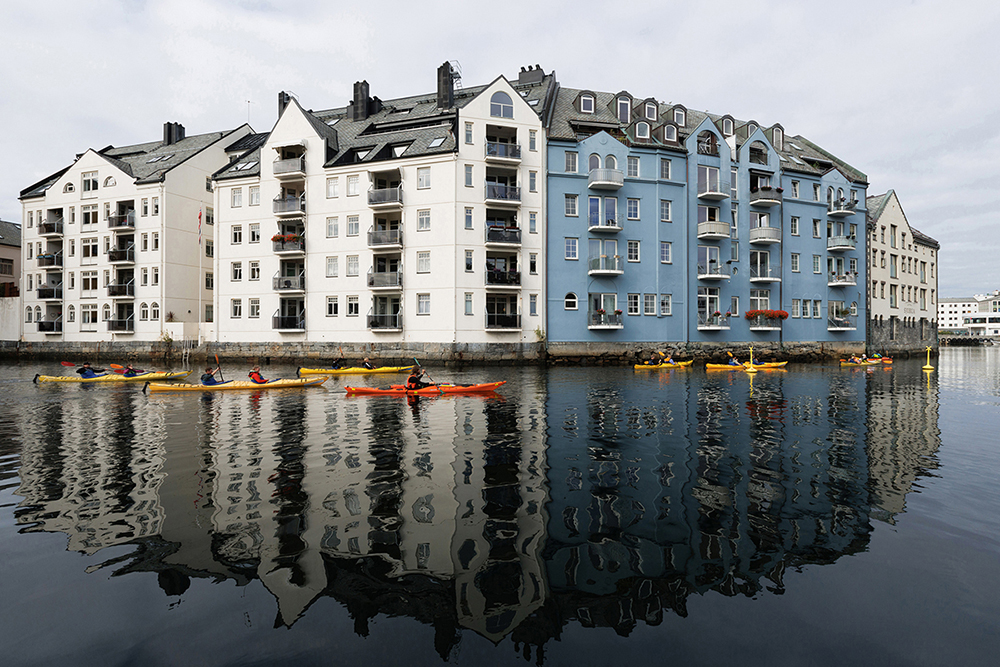 Bunte Kajaks gleiten über ein ruhiges Gewässer und spiegeln unter dem bewölkten Himmel die weißen und blauen Gebäude am Ufer wider.