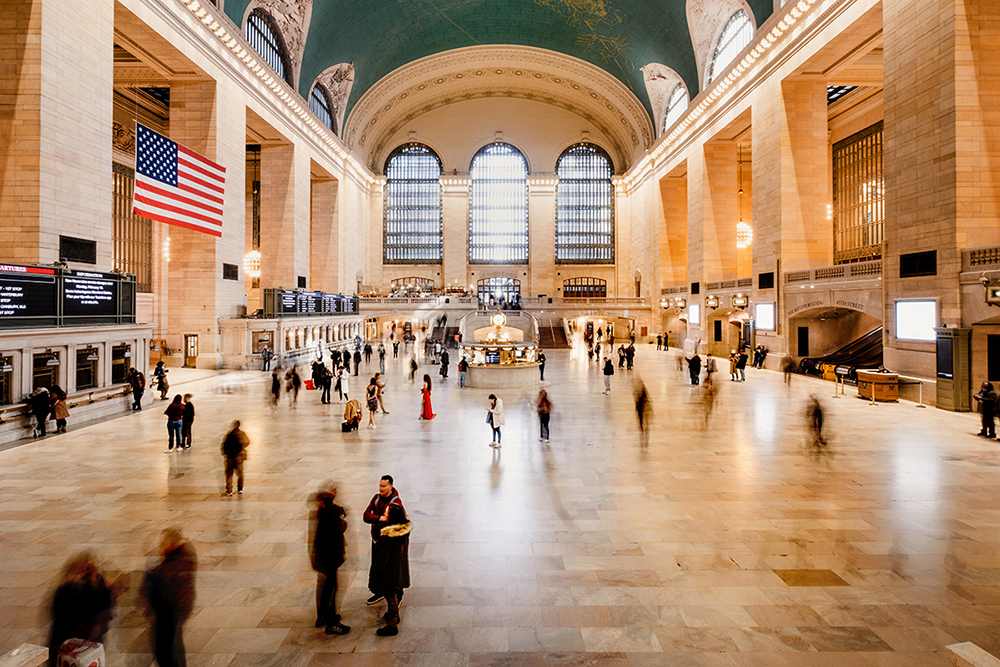Die Haupthalle des Grand Central Terminal mit Menschen, die durch die Halle gehen, einer amerikanischen Flagge, Bogenfenstern und einer hohen Decke.