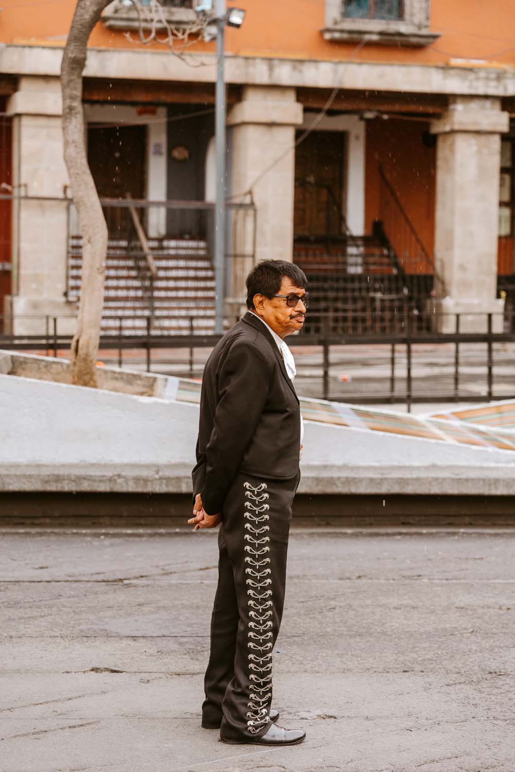 Man in a black charro suit with silver embellishments stands on a street, hands behind his back, with a building in the background.