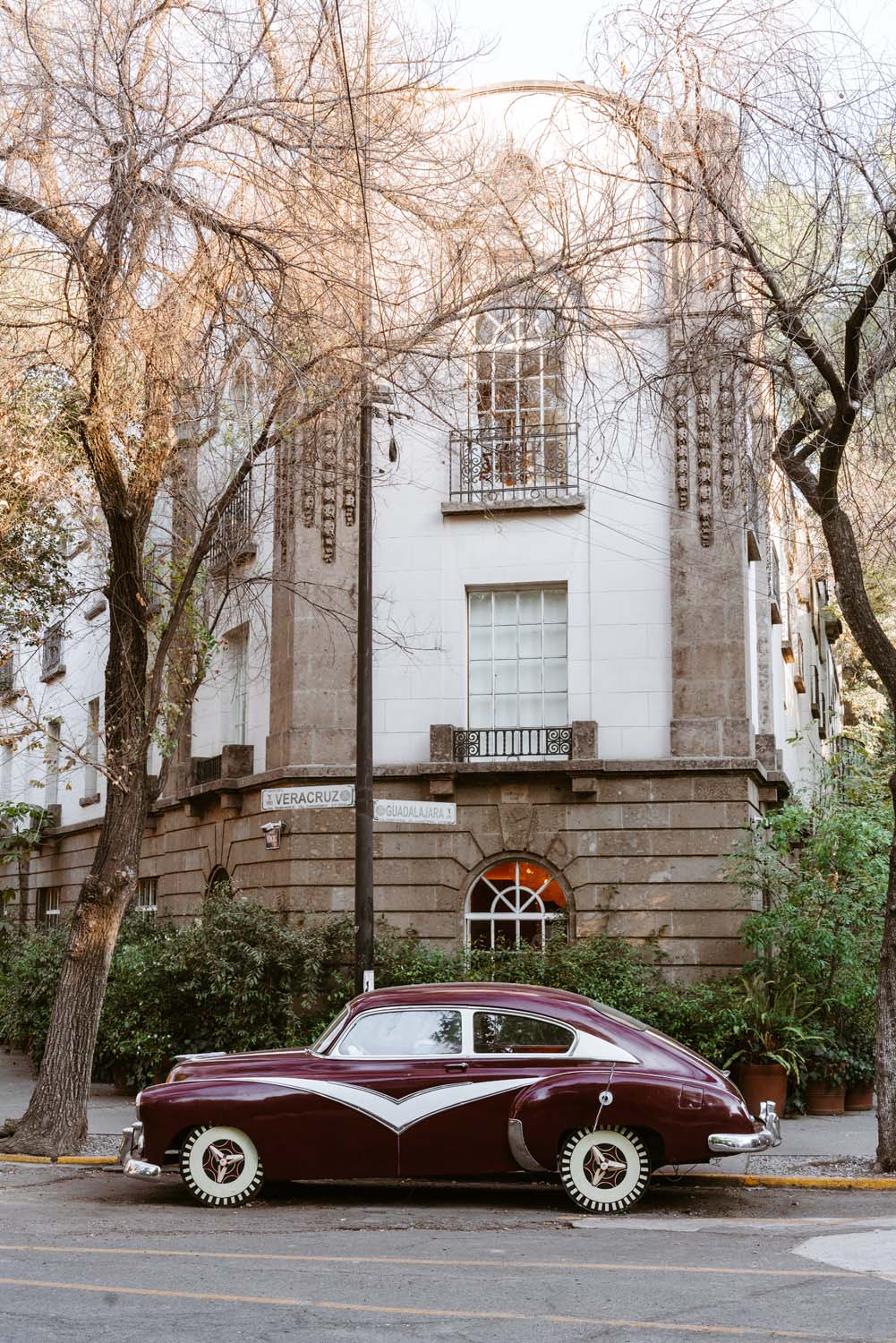 A vintage maroon car parked on a street beside a historic building with ornate architecture and bare trees.