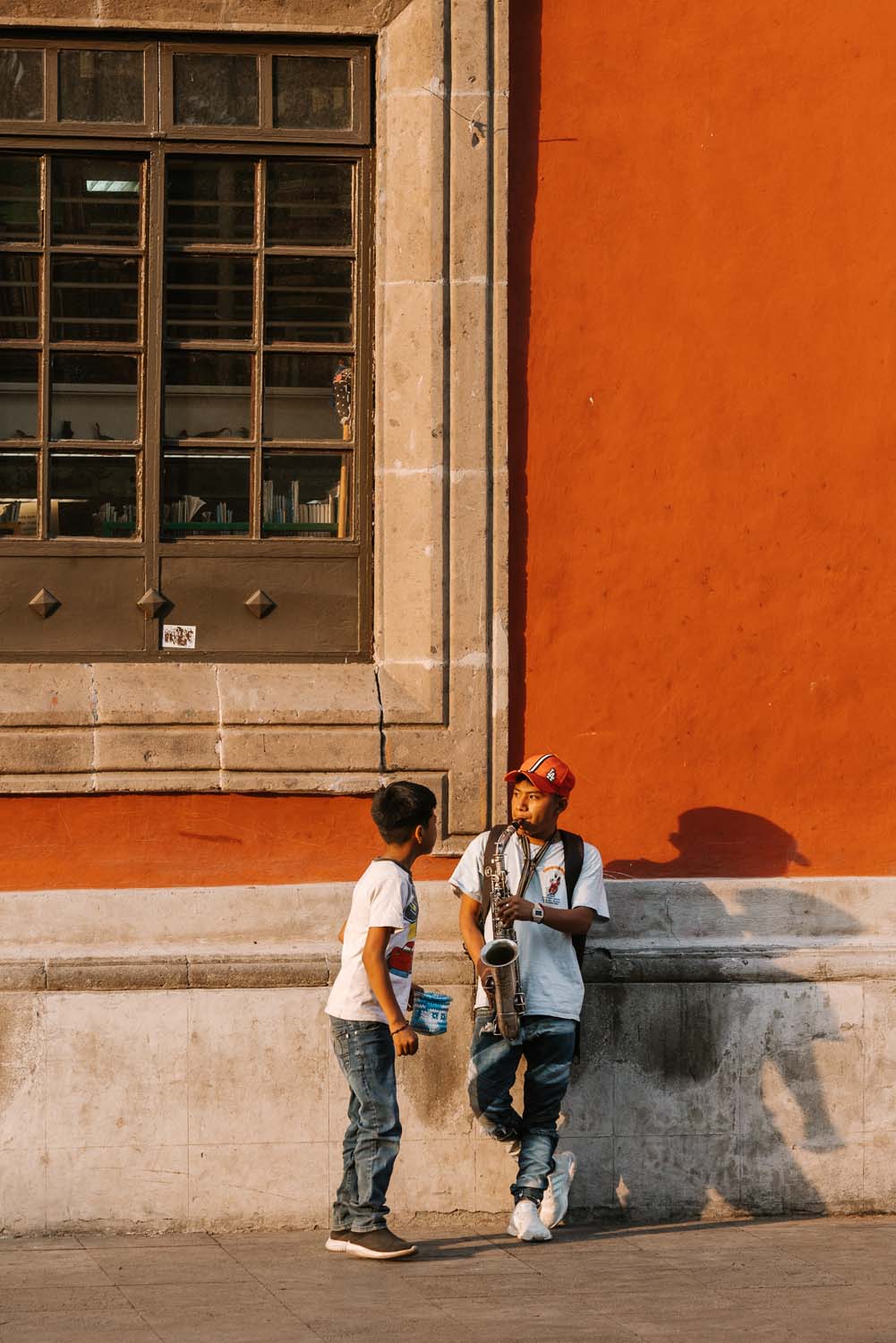Two people converse beside a red wall with a large window. One leans against the wall, while the other stands holding a small container.