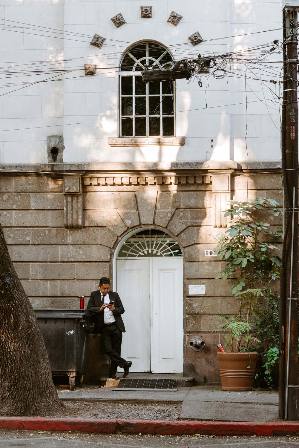 Man in a suit leans against a building, reading a newspaper. A large potted plant and a trash bin are nearby on the sidewalk.