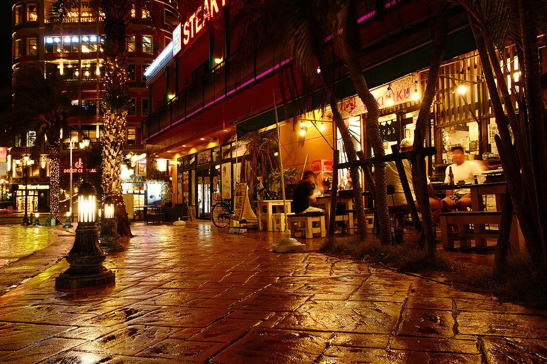 Una strada illuminata di notte, con un ristorante su un lato, tavolini all'aperto e luci decorative che illuminano l'asfalto bagnato.