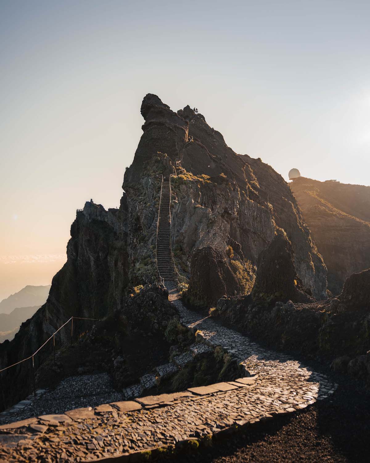 Sunlit mountain path with stone steps leading up a rugged peak, surrounded by rocky terrain and a clear sky in the background.