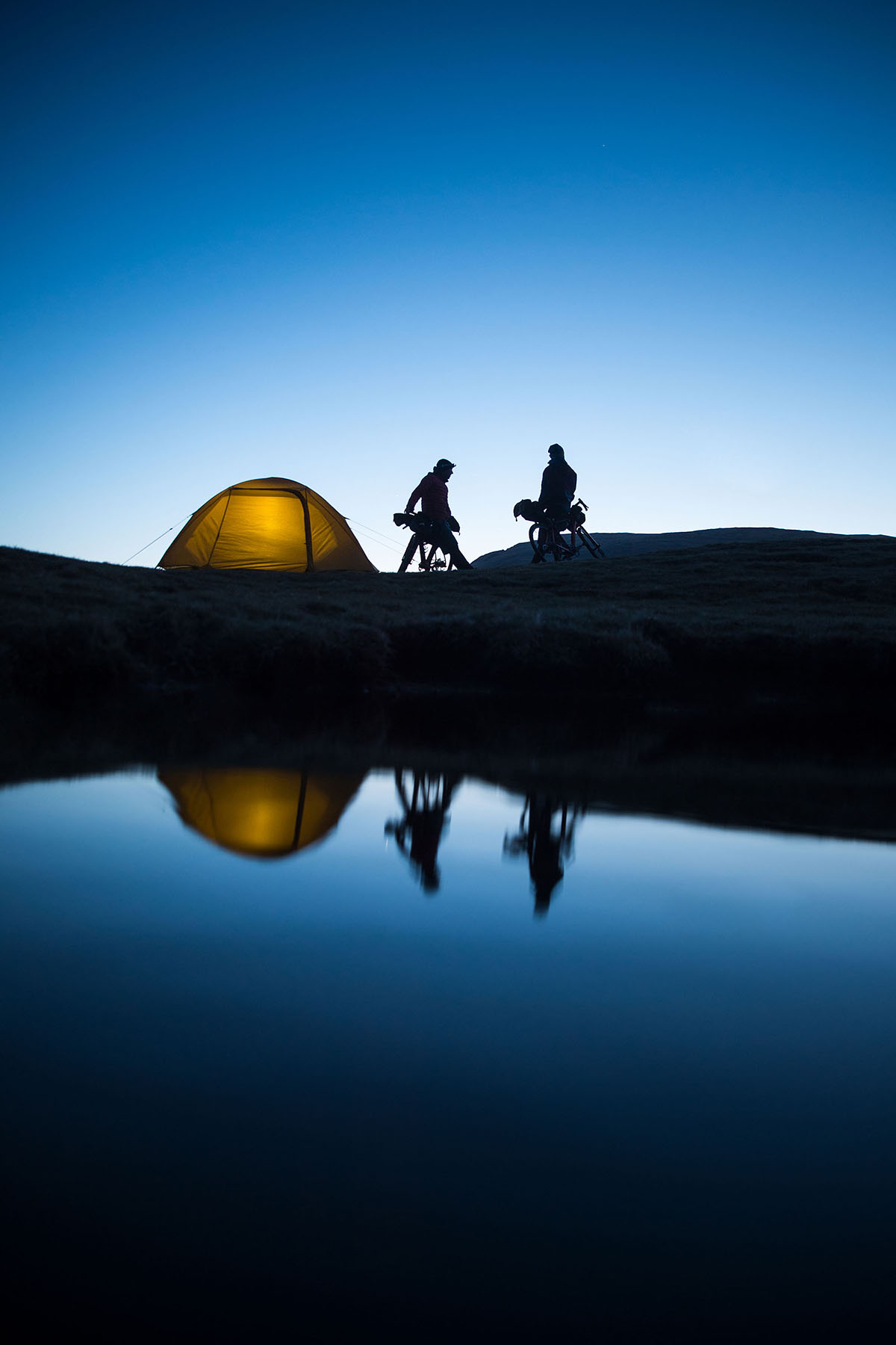 Zwei Radfahrer und ein leuchtendes Zelt zeichnen sich vor dem Abendhimmel ab, ihre Spiegelbilder spiegeln sich in einem ruhigen Gewässer.
