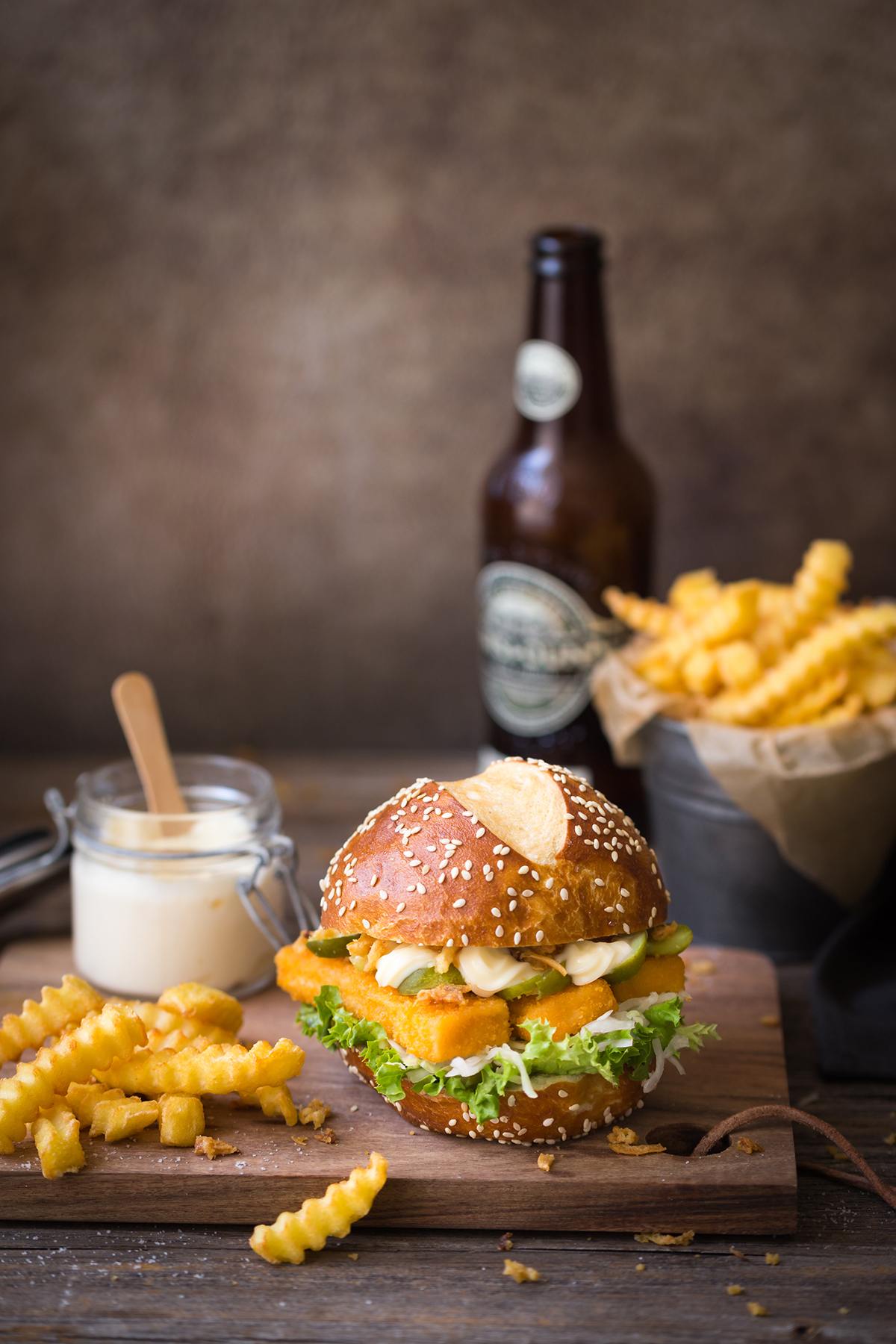 Ein Fischburger mit Salat, Käse und einer Pommes oben drauf, serviert mit welligen Pommes, Mayonnaise und einer Flasche Bier auf einem Holzbrett.
