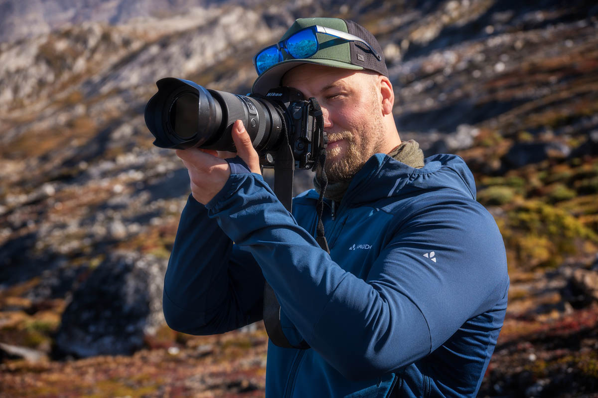 A man in a blue jacket takes a photo with a camera outdoors, wearing a cap with sunglasses on top. Rocky landscape in the background.