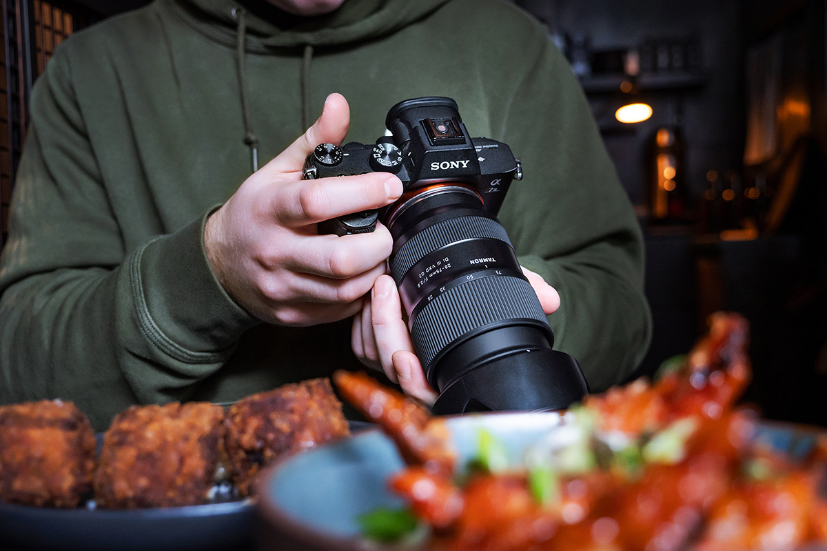 Eine Person in einem grünen Kapuzenpulli fotografiert Essen mit einer Sony-Kamera. Im Vordergrund stehen Teller mit Brathähnchen und anderen Gerichten.