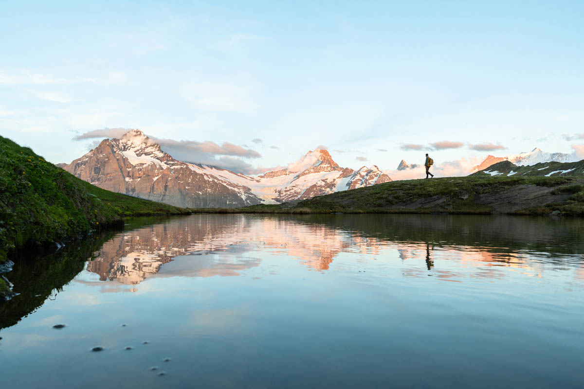 A lone hiker walks near a reflective mountain lake with snow-covered peaks in the background, under a clear blue sky at dawn.