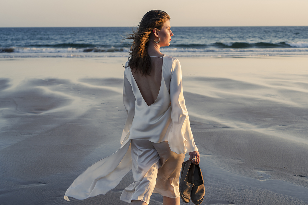 Eine Frau in einem wallenden weißen Kleid läuft barfuß bei Sonnenuntergang am Strand entlang, die Schuhe in der Hand, während im Hintergrund die Wellen sanft an den Strand schlagen.