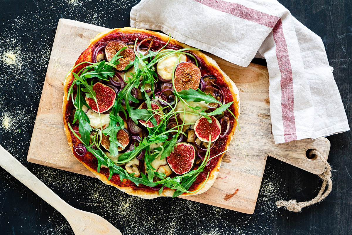 Eine rustikale Pizza mit Feigen, Rucola, Zwiebeln und Käse auf einem Holzbrett, dazu ein Tuch und ein Holzlöffel.