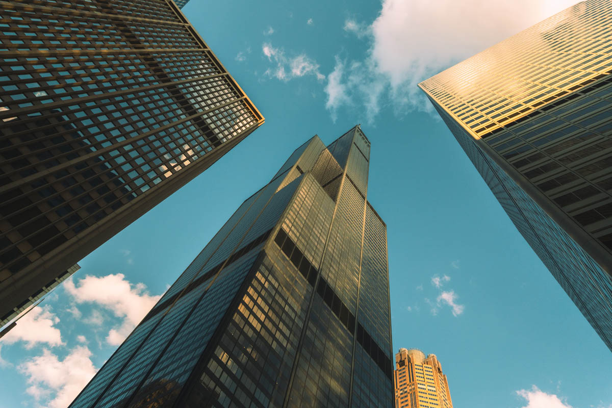Low-angle view of three modern skyscrapers against a clear blue sky with scattered clouds.