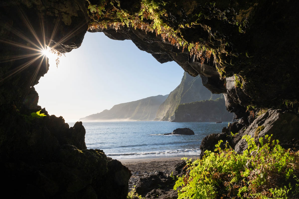 View from inside a cave looking at a beach and ocean, with sun rays peeking through the cave opening and lush greenery foreground.