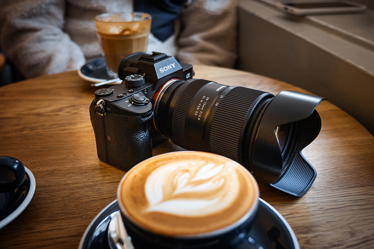 Eine Kamera mit einem großen Objektiv auf einem Holztisch neben einem Latte mit Blattmuster im Schaum und einem weiteren Kaffee im Hintergrund.