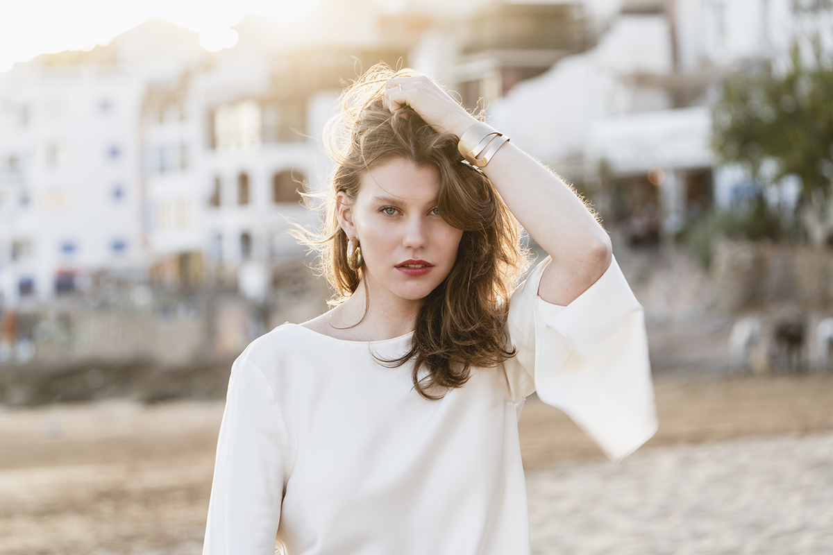 Eine Person mit langen Haaren, die ein weißes Oberteil trägt, steht bei Sonnenuntergang am Strand, im Hintergrund sind verschwommene Gebäude zu sehen.
