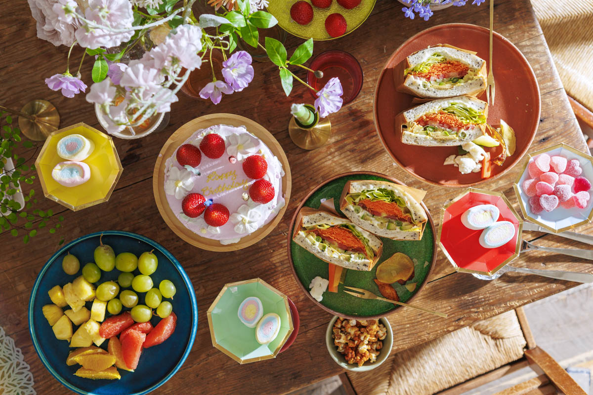 A vibrant spread with a birthday cake, sandwiches, fruit salad, drinks, heart-shaped sweets, popcorn, and pastel flowers on a wooden table.