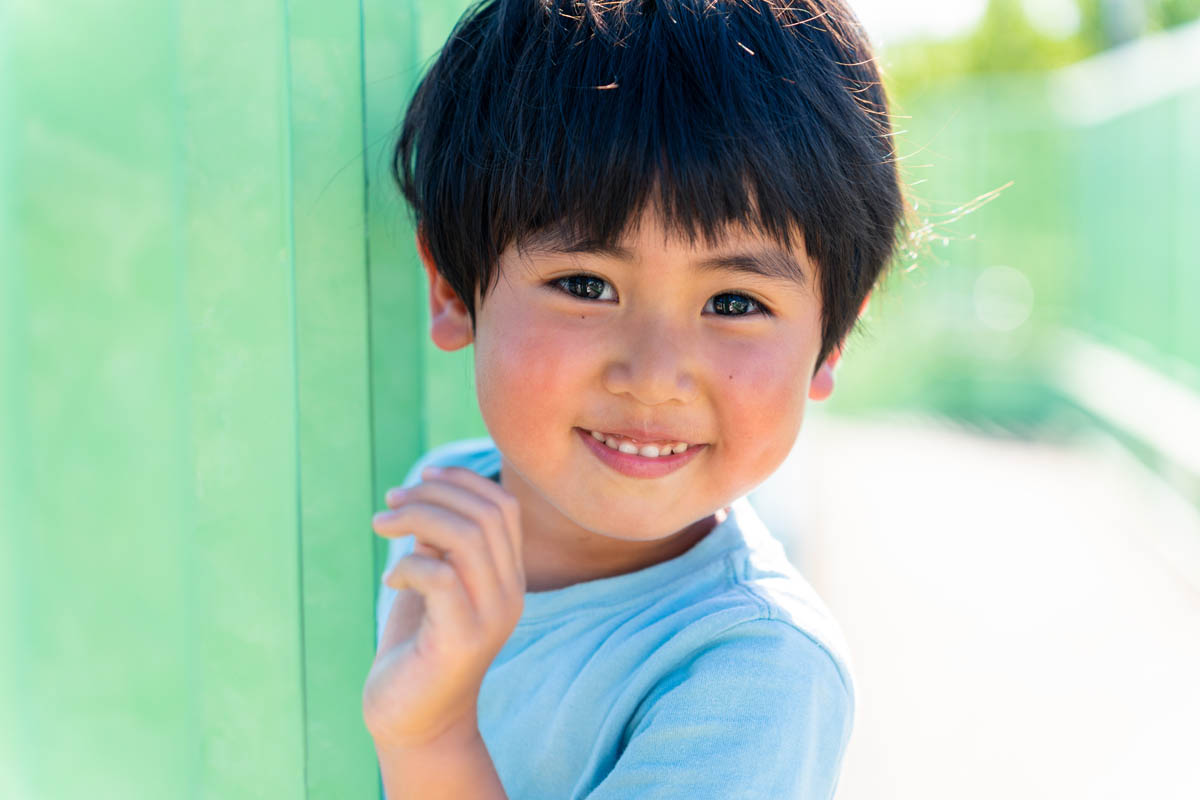 A smiling child in a light blue shirt stands outdoors, leaning against a green fence, with sunlight highlighting their face.