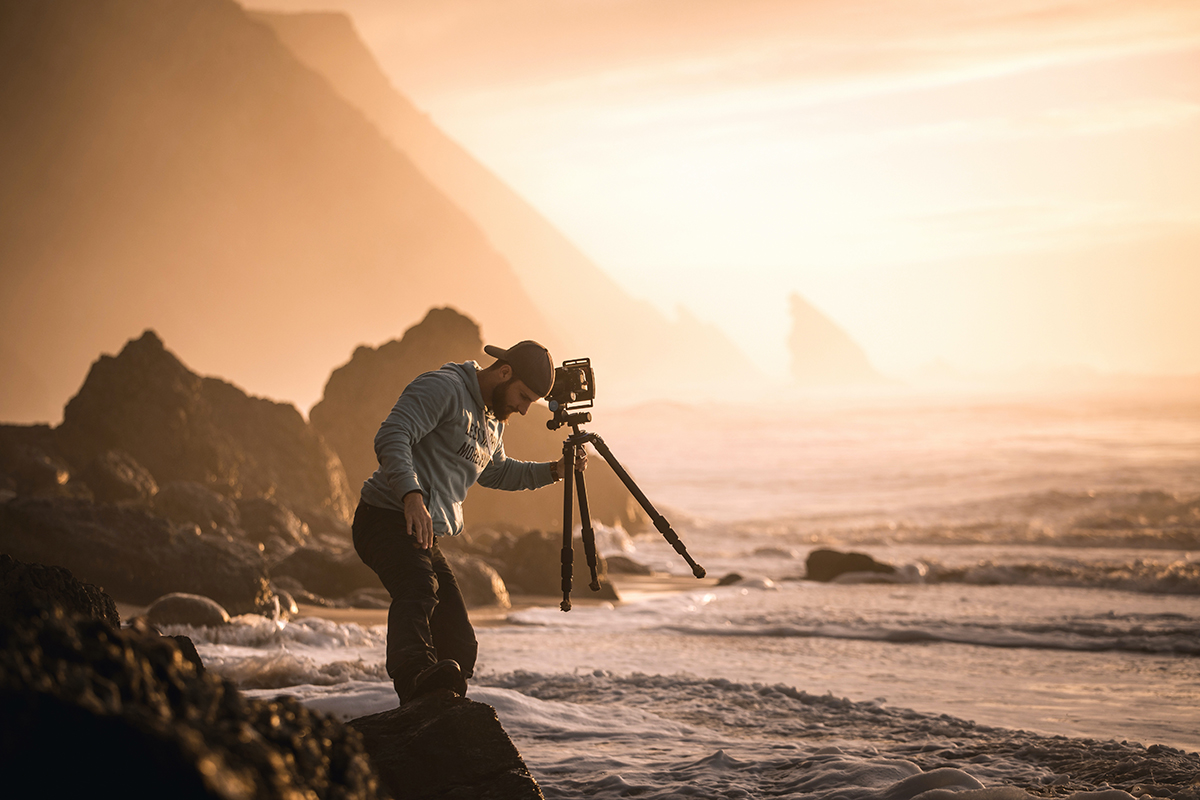 Ein Fotograf an einer felsigen Küste stellt bei Sonnenuntergang seine Kamera auf einem Stativ ein, im Hintergrund sind Wellen und nebelverhangene Klippen zu sehen.