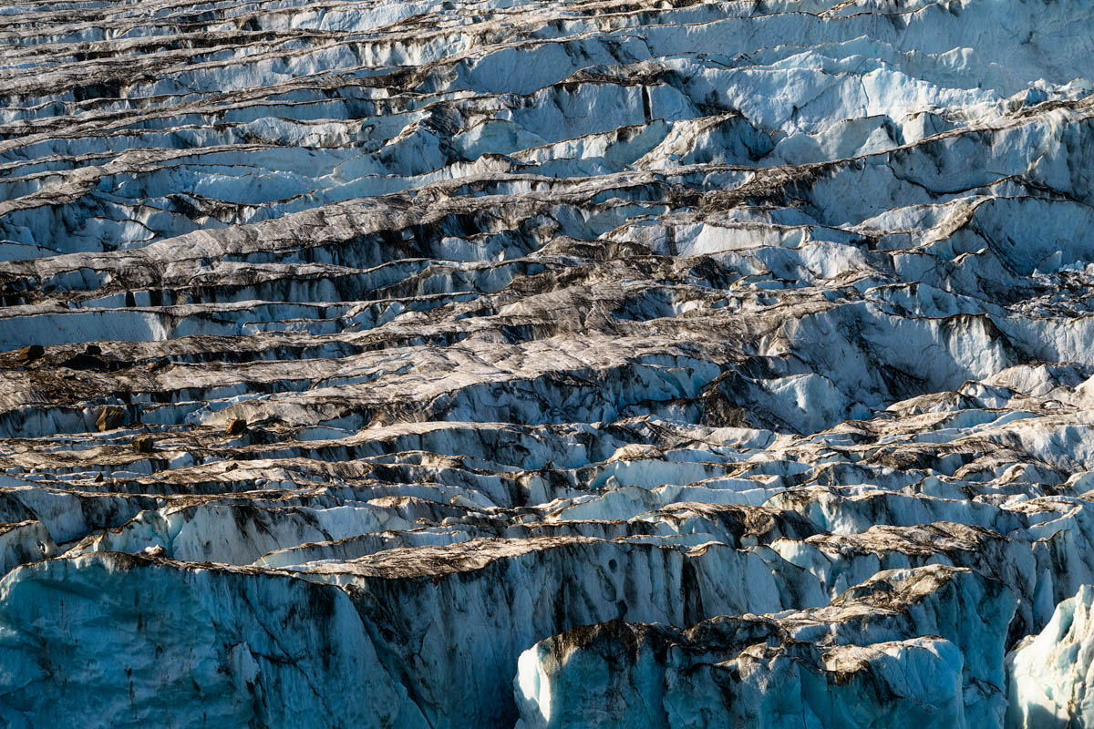 Close-up of a glacier's jagged, blue ice surface with dark sediment lines, creating a striking contrast and rugged texture.