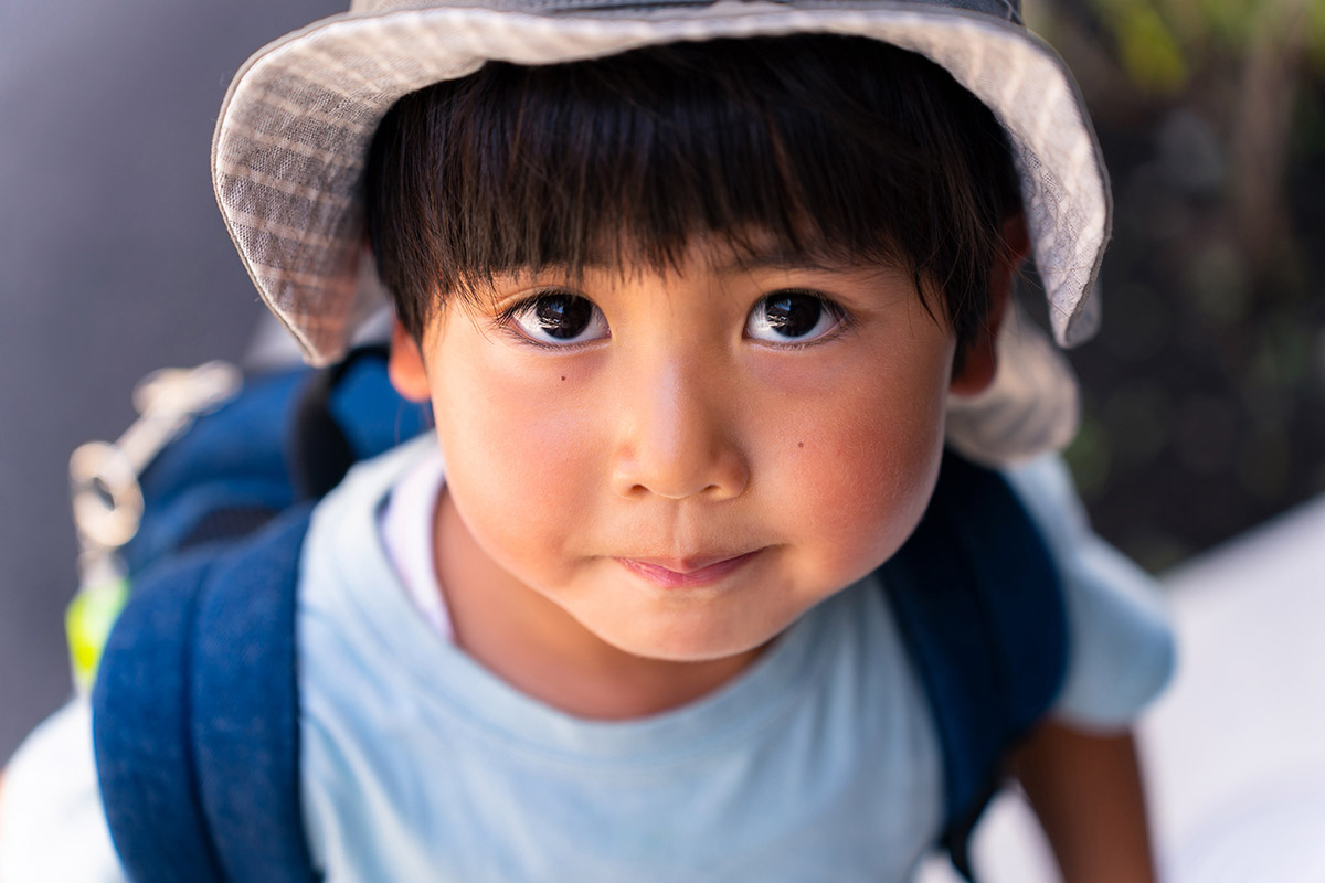 A young child wearing a hat and backpack looks up at the camera with wide eyes and a slight smile.