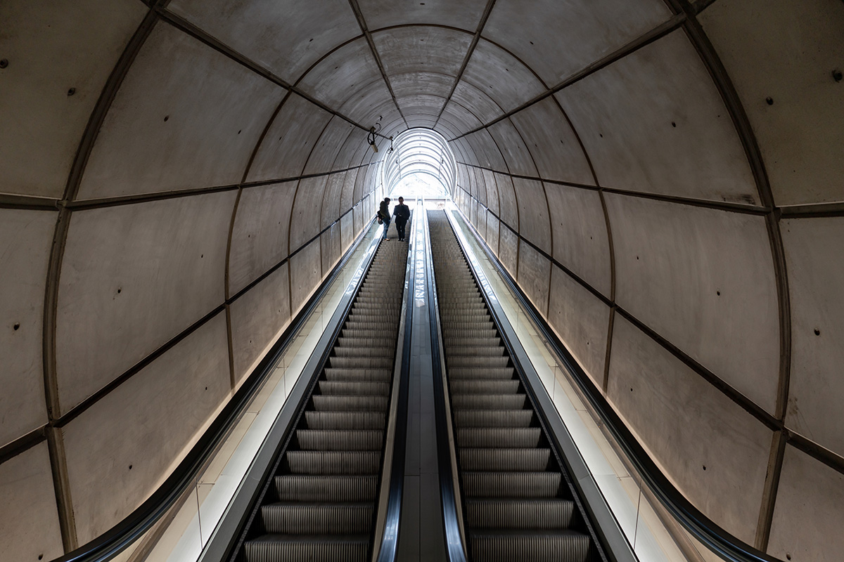 Eine lange Rolltreppe in einem geschwungenen Tunnel, auf der zwei Menschen dem hellen Licht am oberen Ende entgegenfahren, was eine dramatische Perspektive erzeugt.