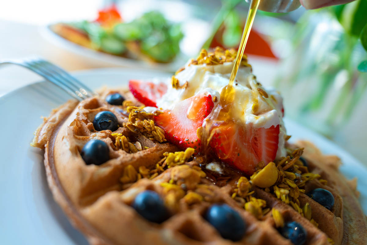 A waffle topped with fresh strawberries, blueberries, granola, whipped cream, and syrup being drizzled.