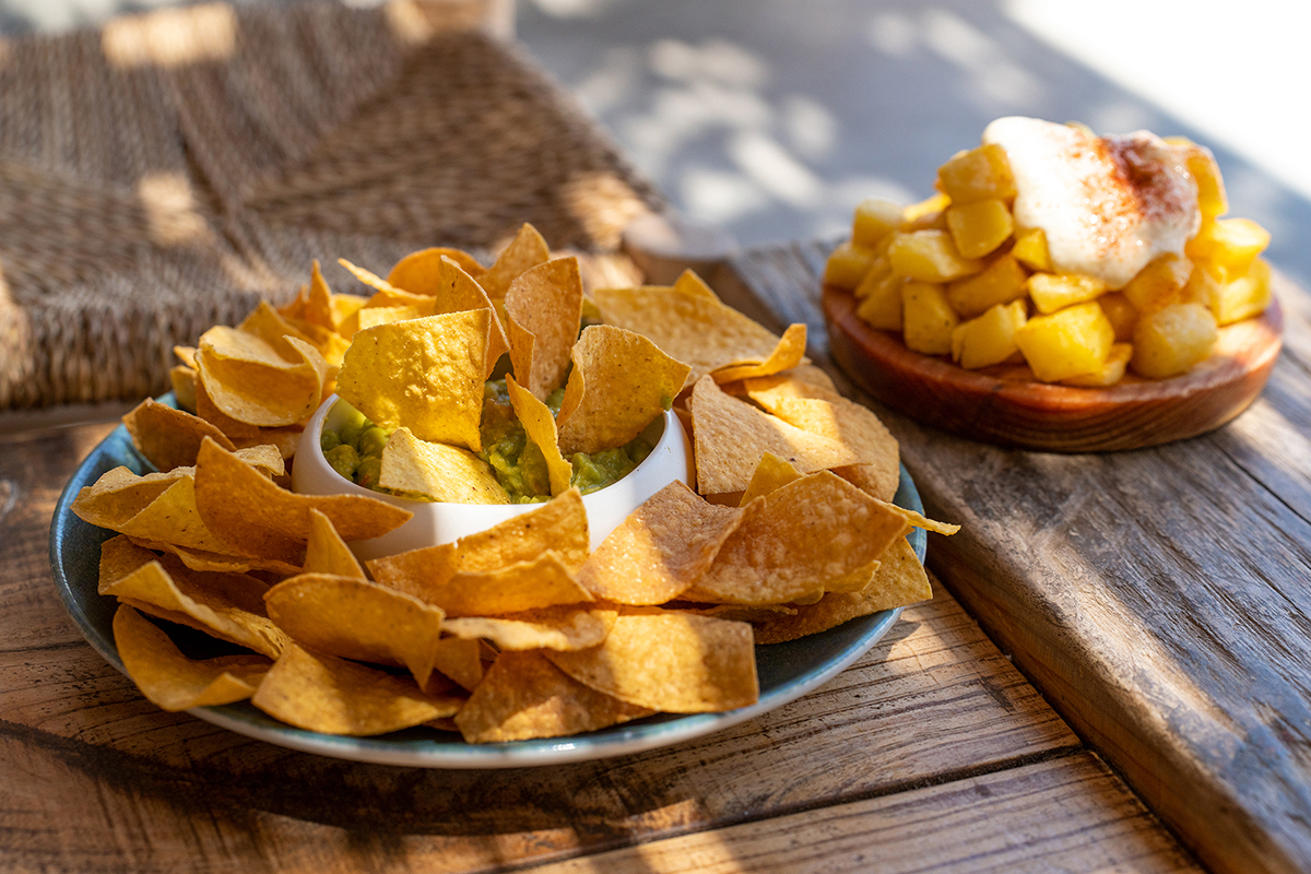 Ein Teller mit Tortilla-Chips, in der Mitte Guacamole, daneben eine Holzschale mit Kartoffelwürfeln, garniert mit einem Klecks weißer Soße, auf einem Holztisch.