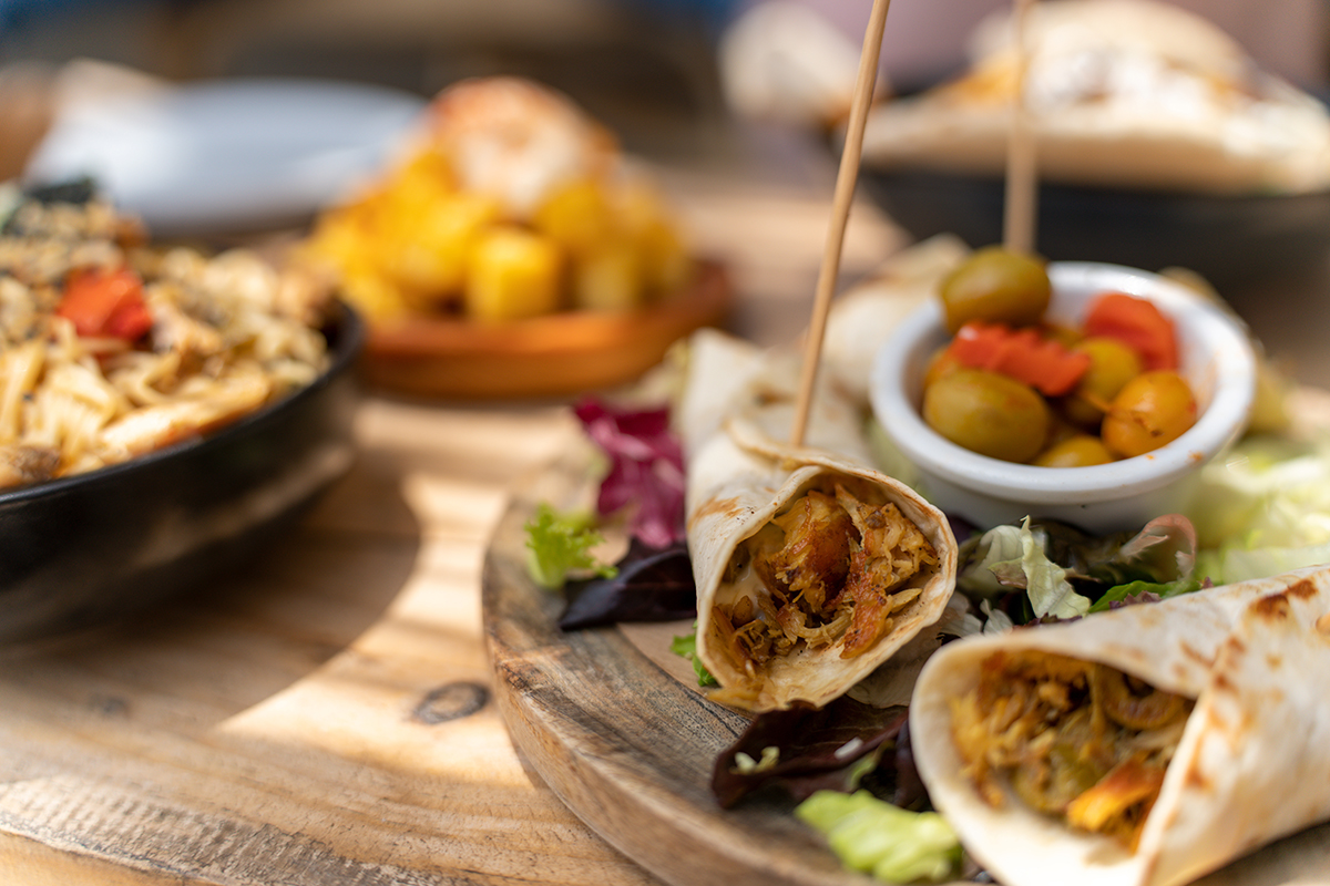 Verschiedene Gerichte auf einer Holzplatte, darunter Wraps gefüllt mit Gemüse, Oliven und gemischtem Salat, im Hintergrund verschwommene Teller.