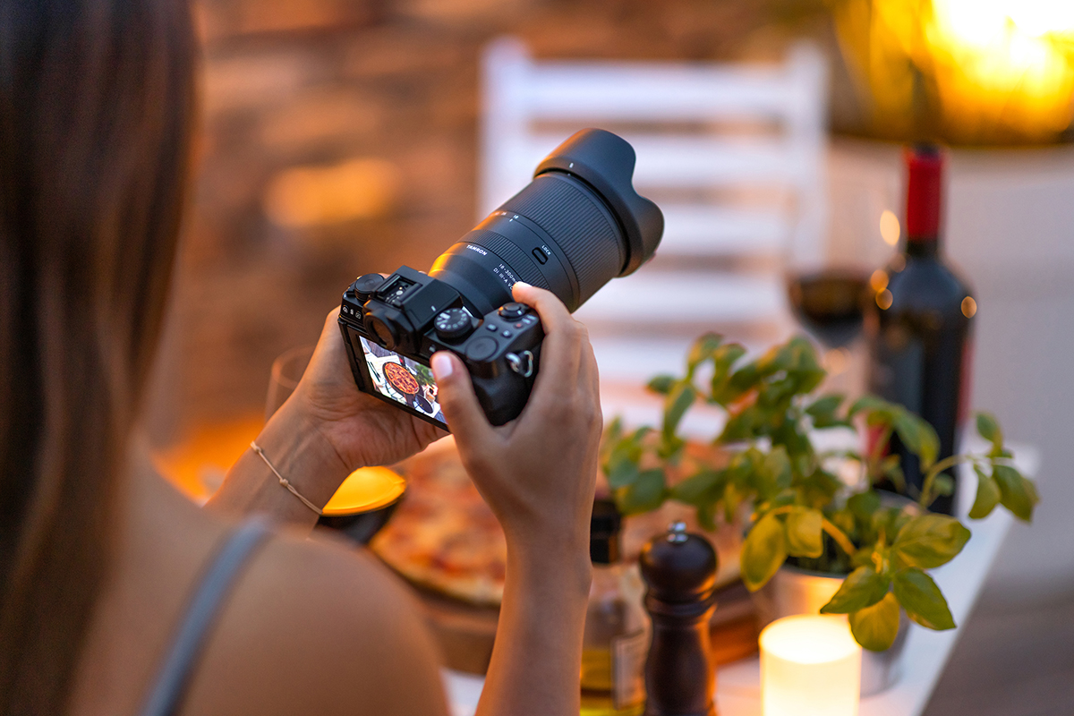 Jemand fotografiert mit einer Kamera Essen auf einem Tisch, umgeben von einer Pflanze, einer Weinflasche, einer Pfeffermühle und einer Kerze, in gemütlicher Beleuchtung.