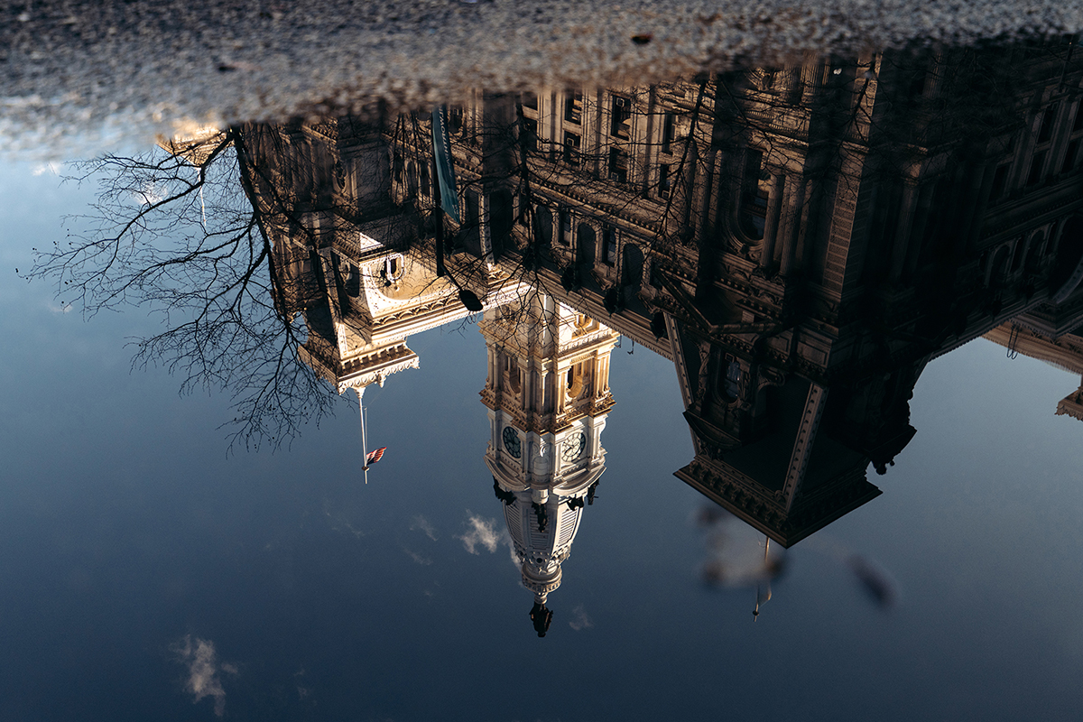 Il riflesso di un edificio riccamente decorato e di alcuni rami in una pozzanghera in una giornata limpida, con il cielo blu sopra.