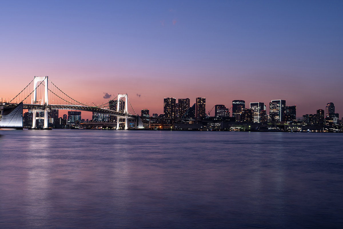 Die Skyline der Stadt in der Abenddämmerung mit einer beleuchteten Brücke über ruhigem Wasser, unter einem leuchtenden, farbenfrohen Himmel.